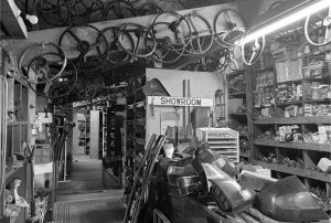 Black and white photo of a vintage car parts showroom filled with shelves of automotive components, steering wheels hanging from the ceiling, and a sign reading 'SHOWROOM' in the center.
