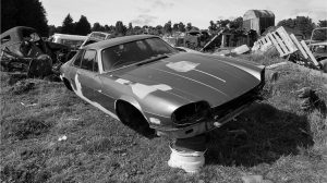 Abandoned vintage car with missing wheels and headlights, propped up on a rim in a grassy junkyard surrounded by other old vehicles and scrap materials.