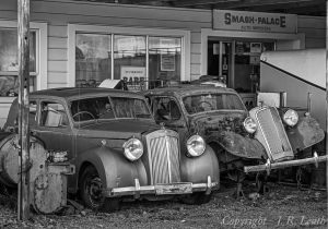 Two vintage cars parked outside Smash Palace Auto Wreckers, one in relatively good condition and the other heavily rusted, with classic grilles and headlights visible.