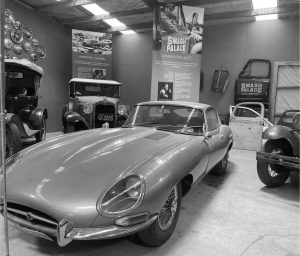 Classic Jaguar E-Type car displayed in a vintage auto showroom, surrounded by older vehicles, hubcaps on the wall, and signage for Smash Palace and Horopito Motors.