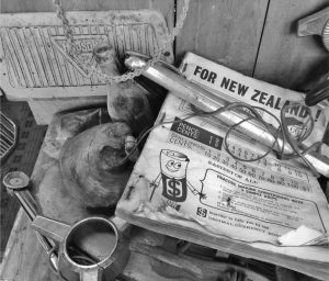 Black and white photo of a cluttered workbench with vintage car parts, tools, and an old New Zealand decimal currency conversion chart featuring a cartoon dollar symbol character.