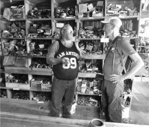 Two men talking inside a vintage auto parts shop, standing in front of shelves filled with assorted car components and boxes.