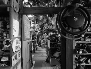 Black and white photo of a vintage auto parts shop interior, showing shelves stacked with car parts, steering wheels, license plates, and hubcaps hanging from the ceiling.